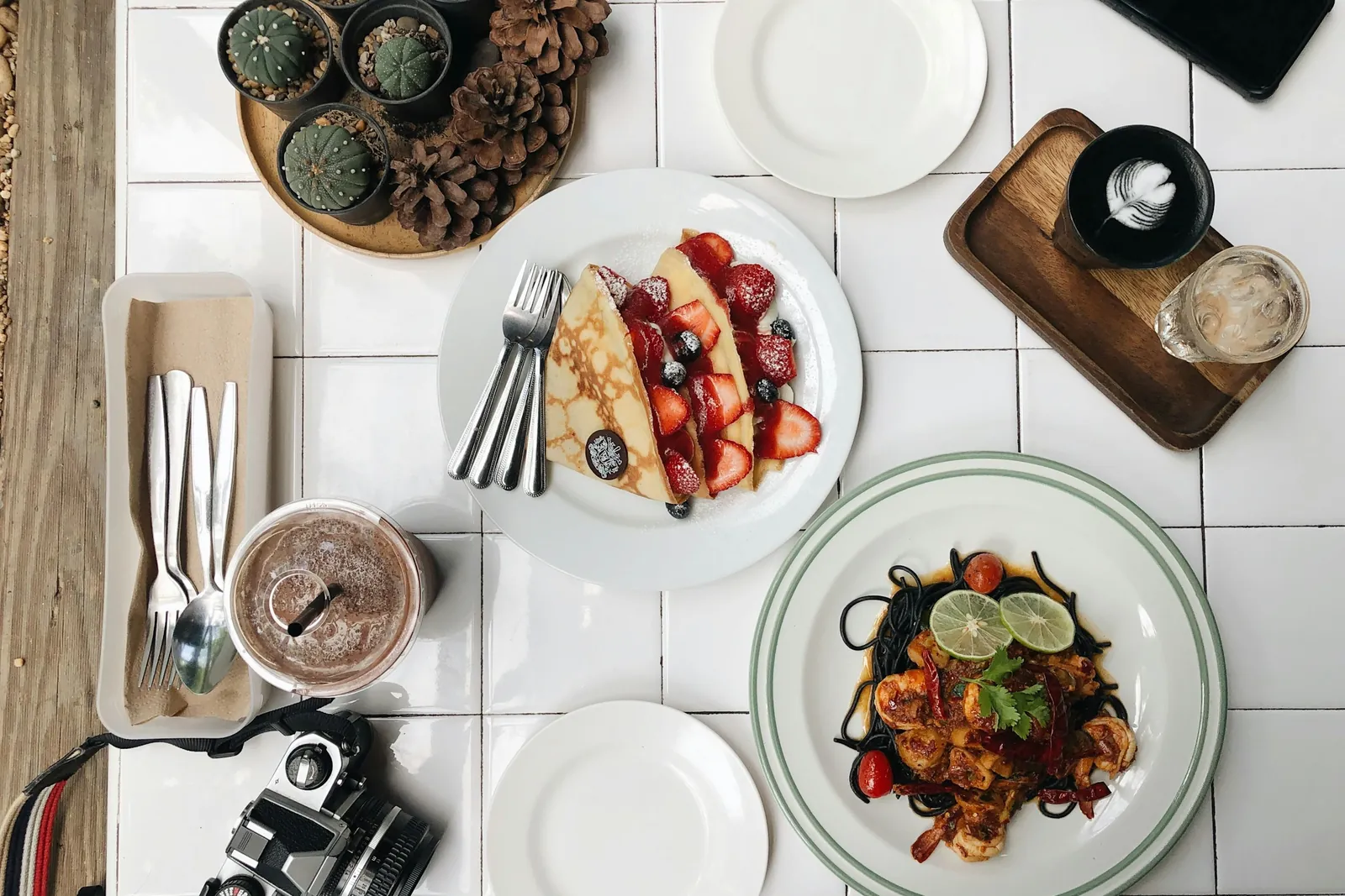 白いタイルの卓を真上から撮った写真。料理の皿のほかに空の皿・カトラリー・カメラ・飲み物が並んでいる